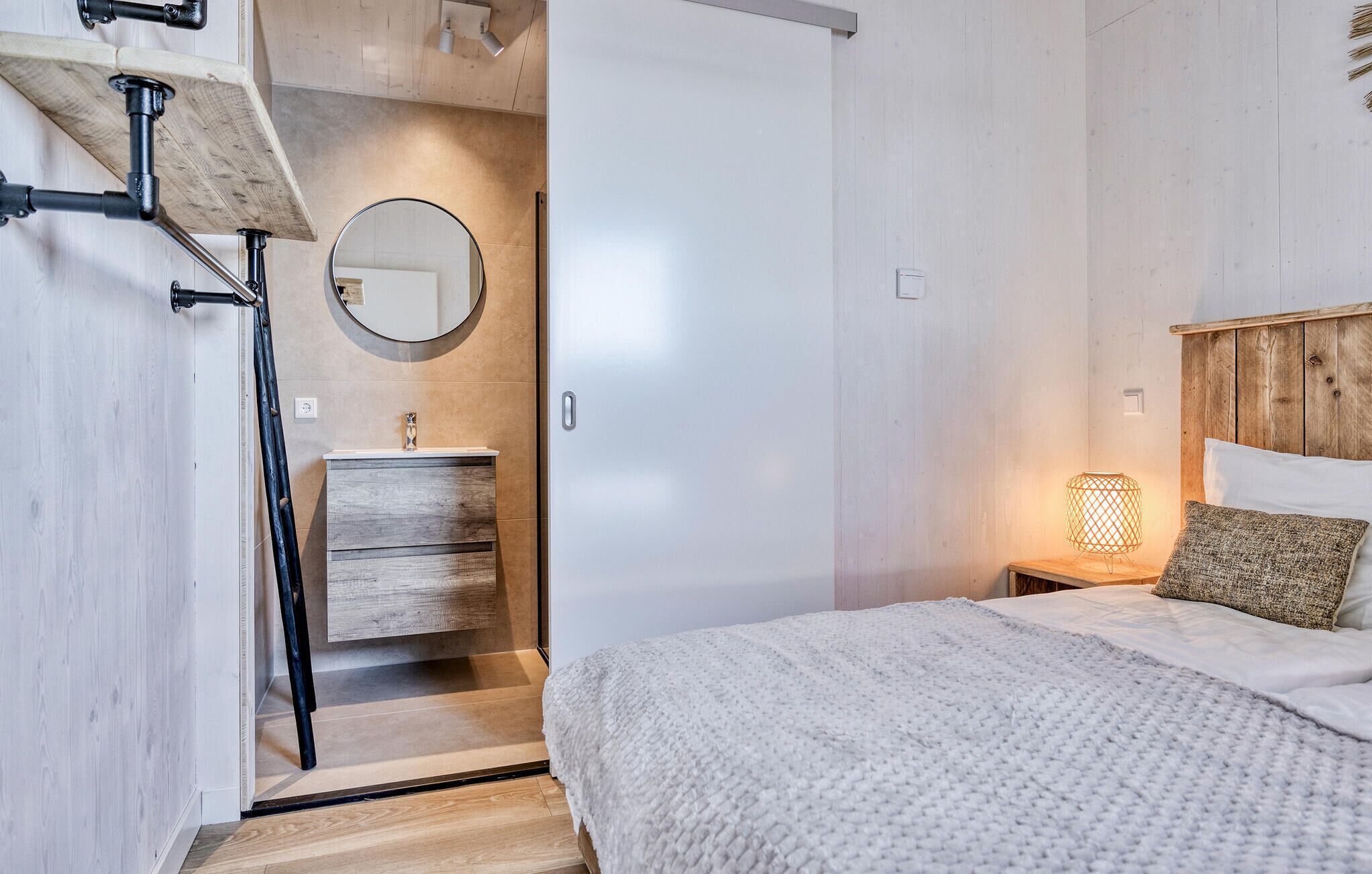 A bedroom with a bed, a bedside table with a lamp, a wooden headboard, a round mirror, and a built-in wardrobe.