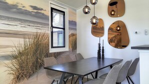 A dining area with a view of the beach through a window, a table with chairs, and wall-mounted spherical light fixtures.