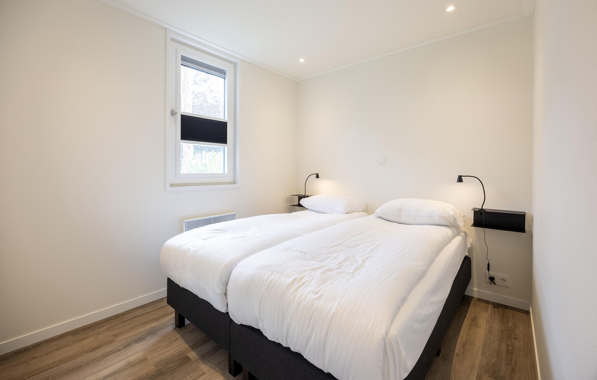 A bedroom with a double bed, two bedside tables, a window with blinds, a wall-mounted heater, and a power outlet.