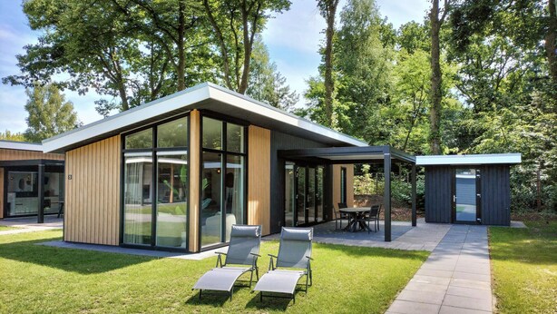 A modern house with large glass windows, a patio with chairs, and a pathway leading to a smaller structure.