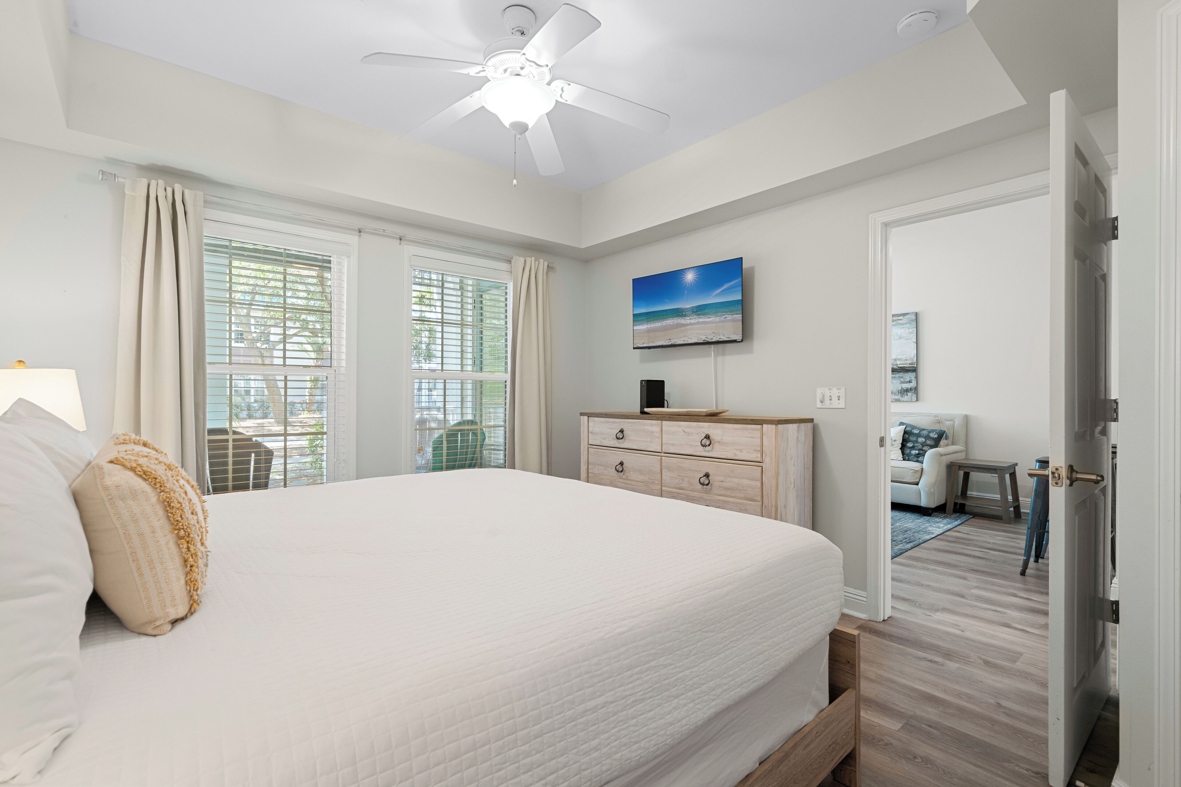A bedroom with a large bed, a ceiling fan, a dresser, a TV, and a door leading to another room.