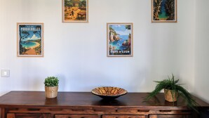 En vägg med fyra inramade affischer som föreställer kustlandskap, ett träbord med en skål och två krukväxter samt ett träskåp med lådor.