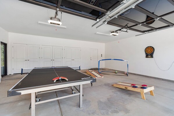A ping pong table, dartboard, and shuffleboard table in a spacious room with white walls and a high ceiling.