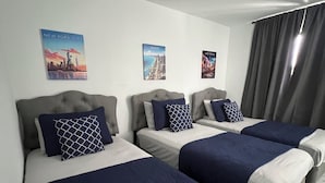 A hotel room with three beds, each with a headboard and pillows. There are wall art depicting New York City, Miami, and Las Vegas.