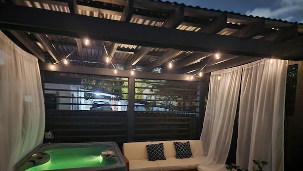 A covered outdoor seating area with a hot tub, a sofa with patterned cushions, and a wooden table.