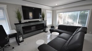 A modern living room with a black leather sofa, a flat-screen TV mounted above a fireplace, and a large window with blinds.