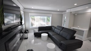 A modern living room with a black leather sectional sofa, white round coffee tables, and a flat-screen TV mounted on the wall.