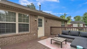 Una casa de ladrillo con puerta principal, un porche con terraza de madera, un sofá negro y una pequeña mesa.