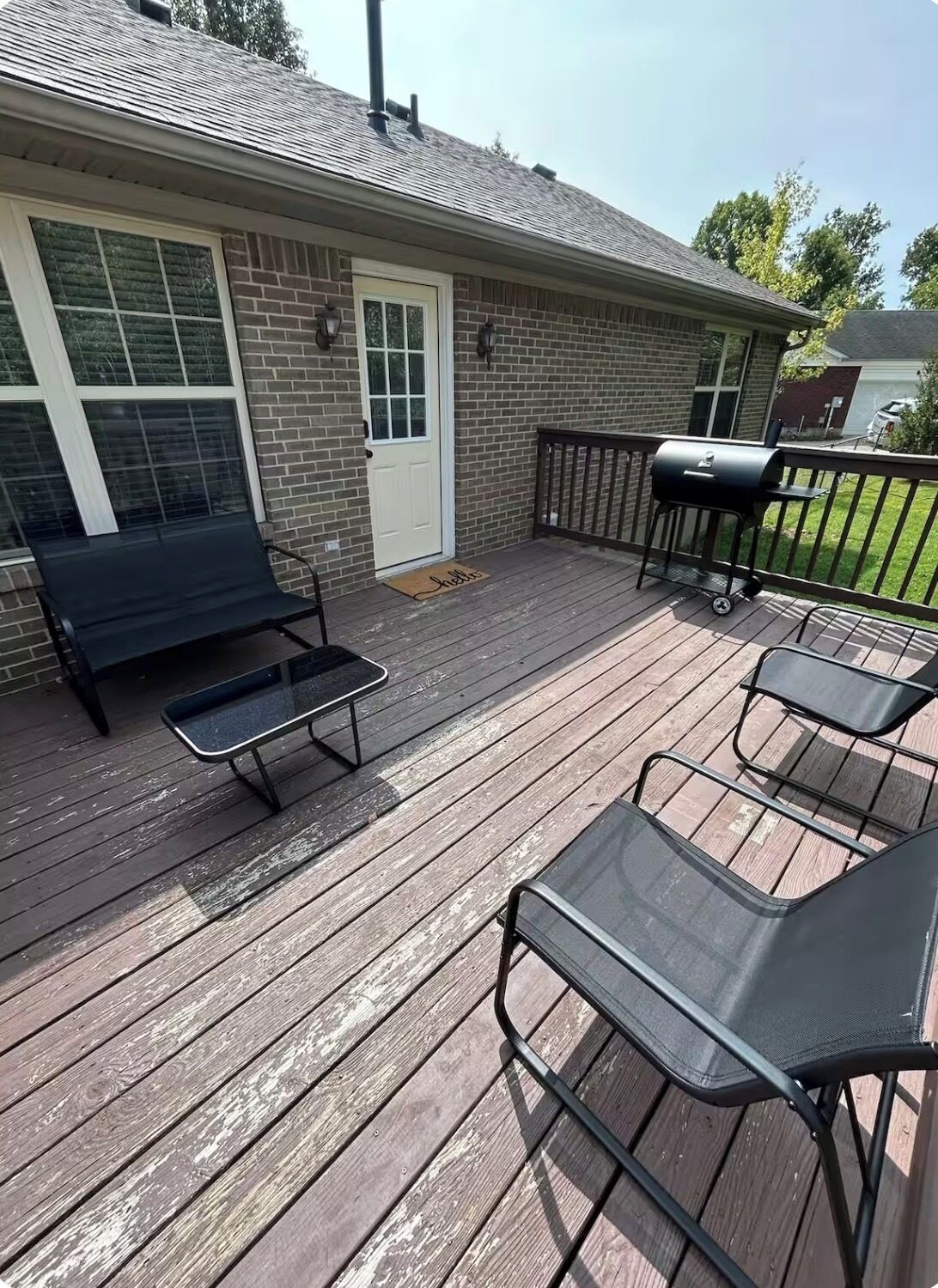 Une terrasse en bois avec des meubles d’extérieur, un barbecue et une maison en briques avec une porte blanche.