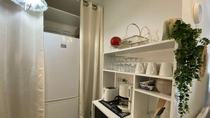 A compact kitchen with a refrigerator, a coffee machine, and shelves with glassware and bowls.