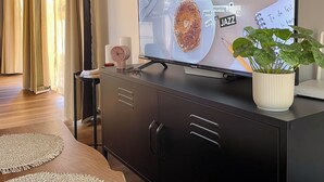 Een moderne woonkamer met een flatscreen televisie op een zwarte kast, een bijzettafeltje met een geweven onderzetter, een potplant en een lijst met kunstwerk aan de muur.