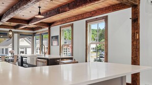 A spacious kitchen with a large island, wooden beams on the ceiling, and large windows offering views of the surrounding area. - Quintessential Vermont Charm in the Heart of Stowe Village (Stowe)