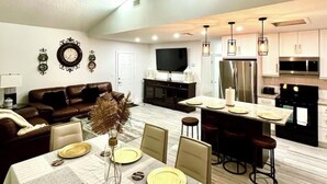 A spacious living area with a dining table set for four, a kitchen with modern appliances, and a ceiling fan.