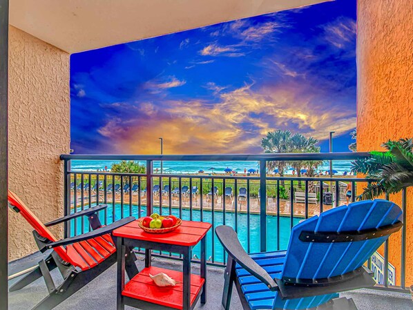 A balcony with a pool, red and blue chairs, a table with fruit, and a mural of a beach scene.
