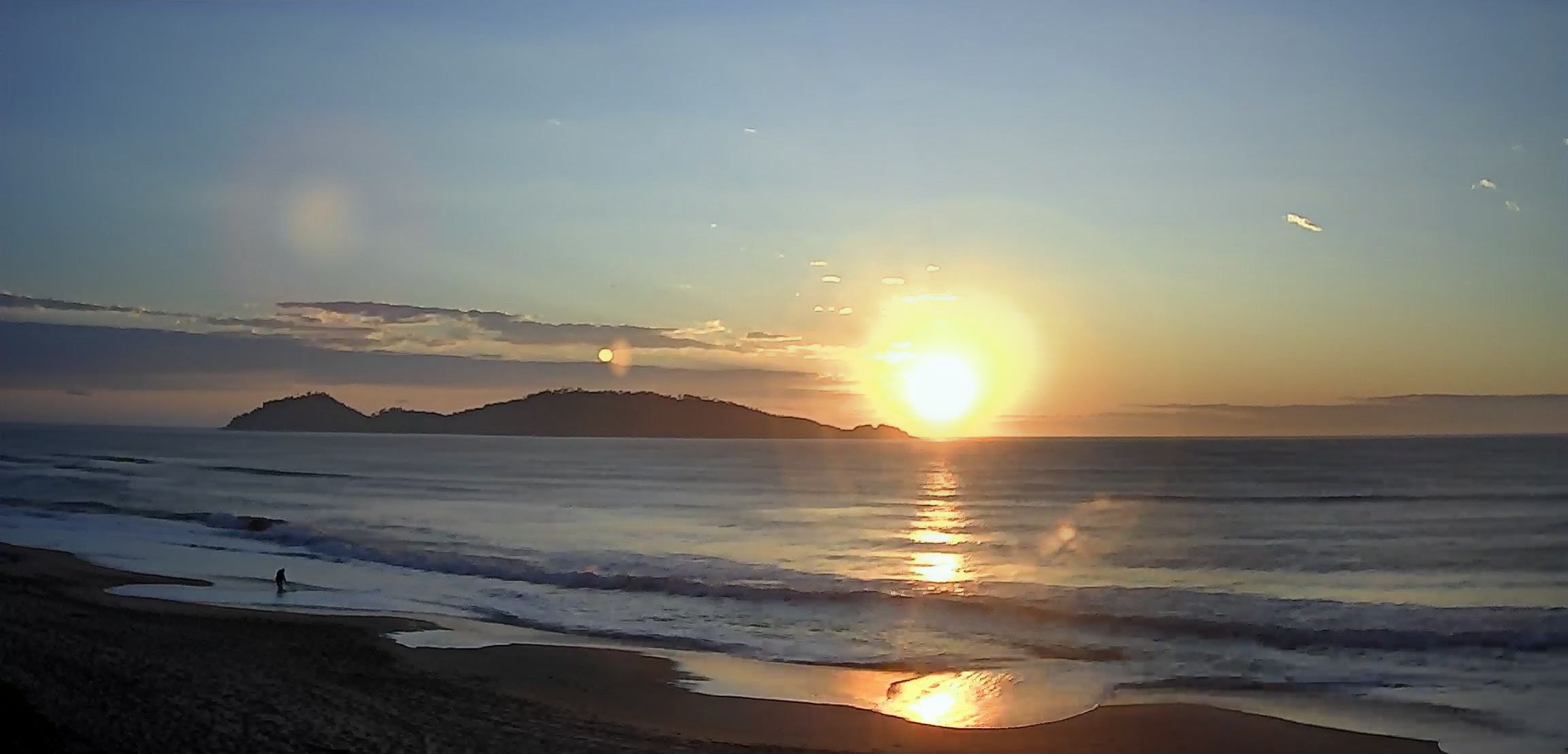 Uma cena de praia com o sol se pondo sobre o oceano, uma montanha ao longe e uma pessoa caminhando pela costa.