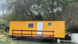 A small, yellow, single-story building with a white door and two windows, situated in a grassy area with trees in the background.