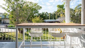 Un balcón con una mesa de madera y sillas blancas, con vistas a un jardín con varios árboles y una casa al fondo.