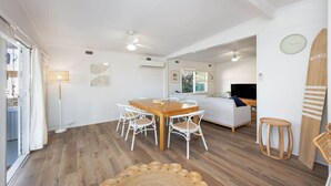 Un luminoso espacio de sala con planta abierta, que incluye una mesa de comedor de madera, sillas blancas, un sofá y un televisor.