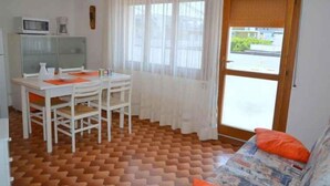 Habitación compacta con mesa y sillas de comedor, un sofá con cojines, una ventana con cortinas translúcidas y una puerta que da al exterior.