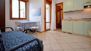 Una habitación pequeña y funcional con cama, una mesa con sillas, kitchenette con armarios verdes y ventana con cortinas.