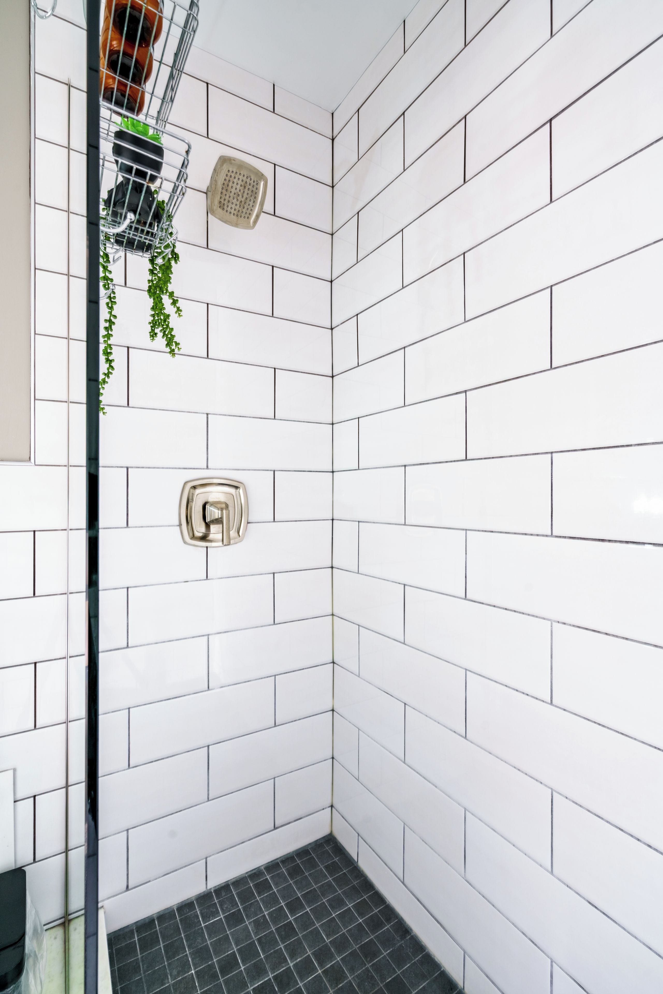 A shower area with white tiled walls, a black tiled floor, a showerhead, and a small potted plant.