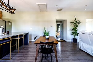 A modern kitchen with a wooden countertop and bar seating, a dining area with a wooden table and chairs, and a living area with a sofa and a potted plant.