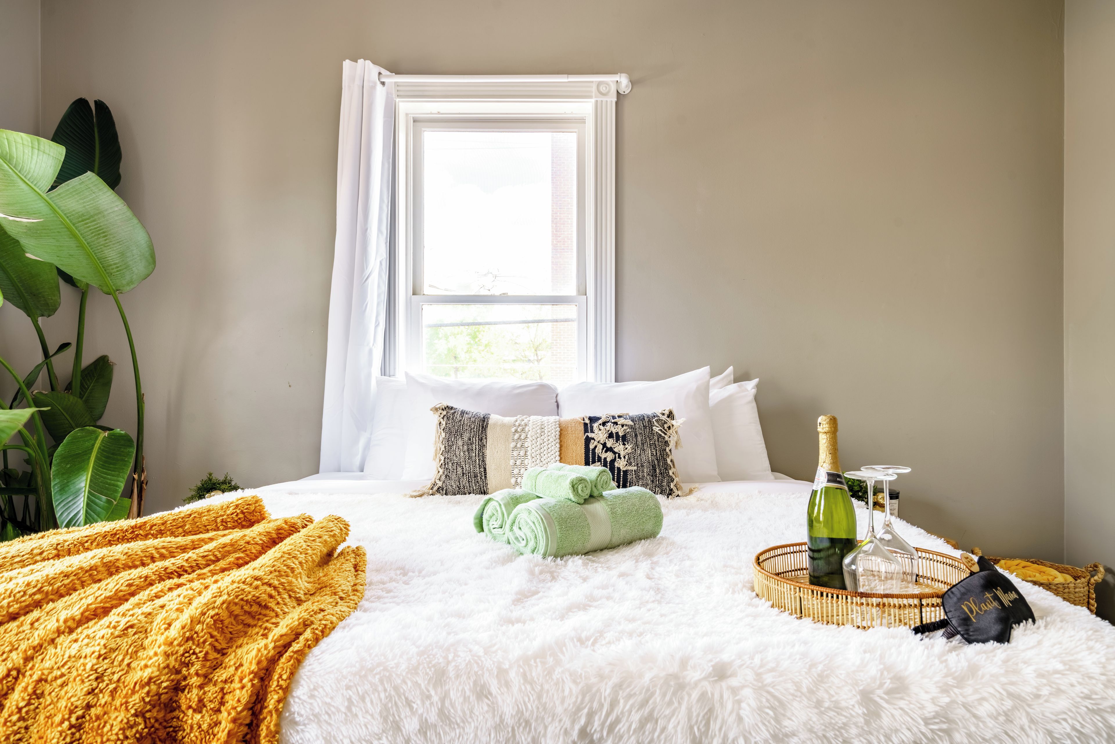 A neatly made bed with a fluffy white duvet, a yellow throw blanket, and a green towel. There is a bottle of champagne and a glass on a woven tray.