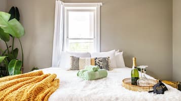 A neatly made bed with a fluffy white duvet, a yellow throw blanket, and a green towel. There is a bottle of champagne and a glass on a woven tray.