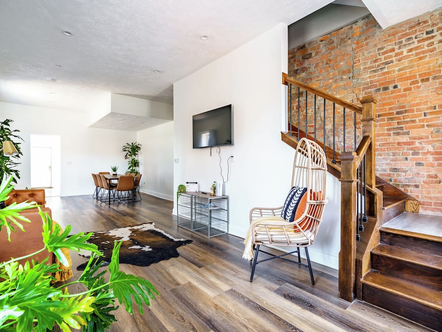 A spacious living area with a wooden staircase, a dining table with chairs, and a TV mounted on the wall.