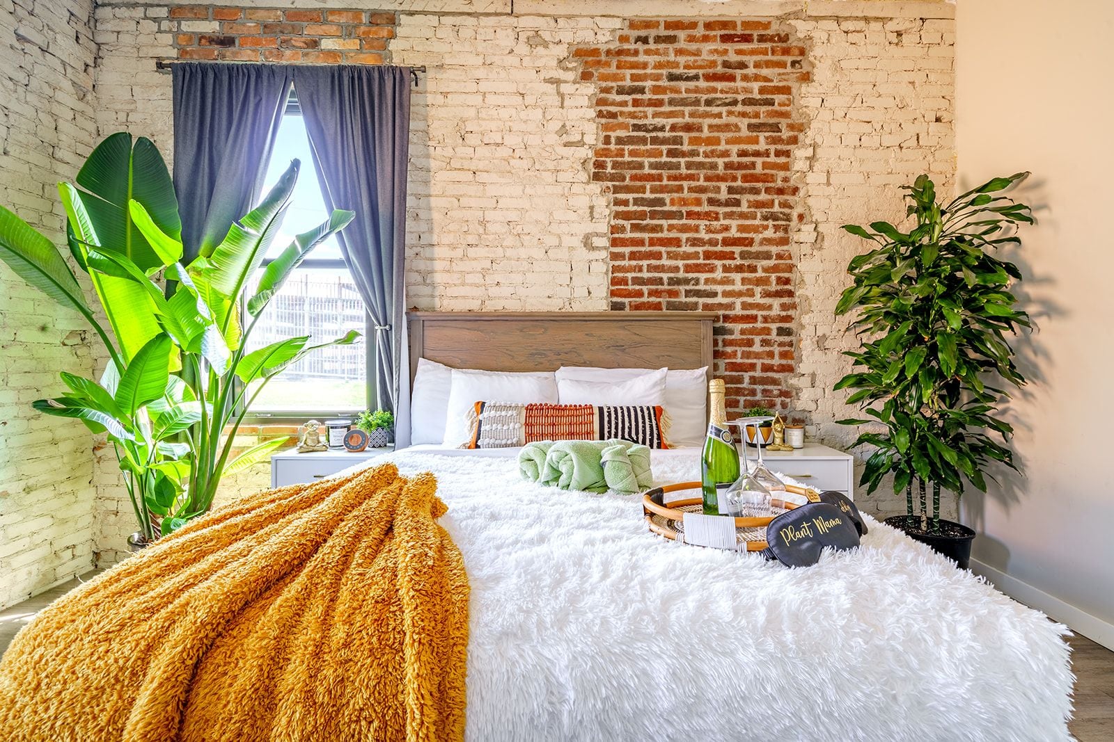 Ein Schlafzimmer mit Backsteinwand, einem Bett mit weißer Bettwäsche, einer großen Pflanze und einem Kissen mit Pflanzenmotiv.