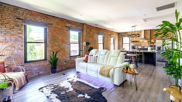 Ein geräumiger Wohnbereich mit einem weißen Sofa, einer rustikalen Backsteinwand und einer Küche im Hintergrund.