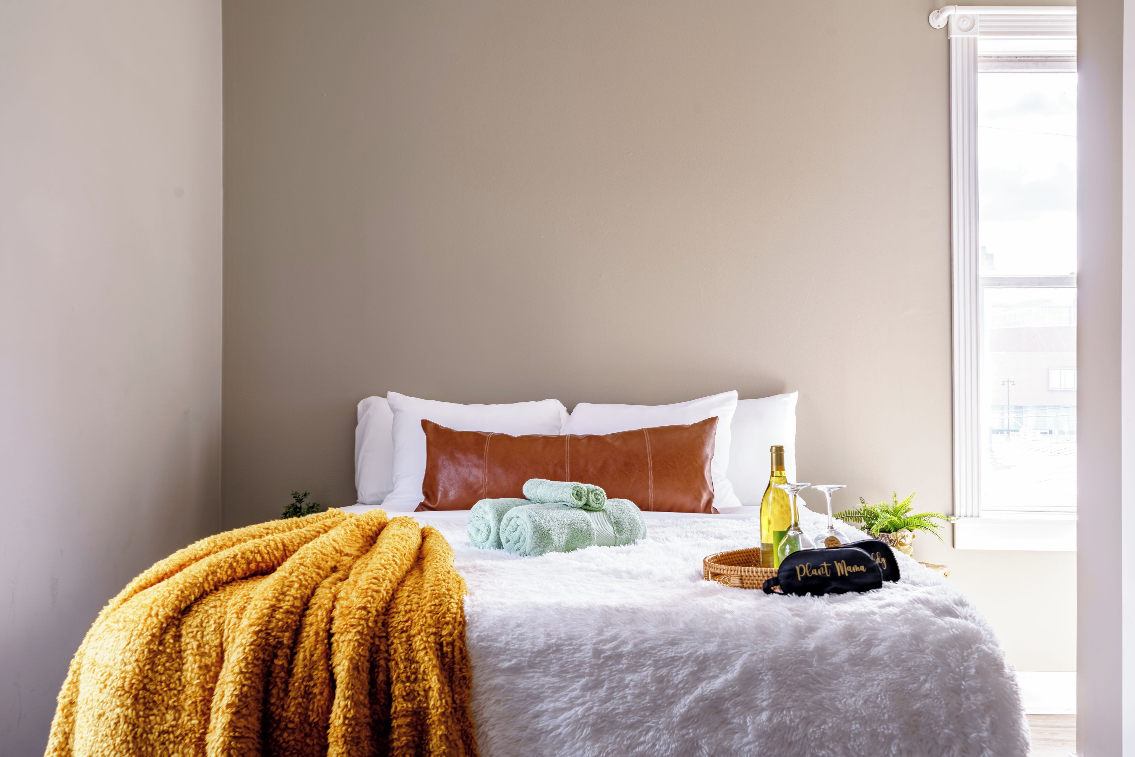 Cama bem arrumada com travesseiros brancos e castanhos, um cobertor amarelo e uma garrafa de vinho ao lado.