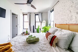 Une chambre à coucher avec un lit, un ventilateur de plafond, une fenêtre avec des rideaux, une plante et une tête de lit en bois.