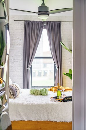 A bedroom with a white fluffy bed, a ceiling fan, a window with curtains, and a view of the outside. - Jungle House Downtown Columbus – Modern Boutique Suites (Columbus)