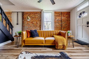 A modern living room with a yellow sofa, a wooden floor, a brick wall, and a staircase.