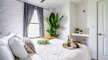 A bedroom with a white brick wall, a large window with curtains, a fluffy white bed, a potted plant, and a bottle of champagne on the bed.