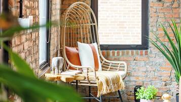 A room with a brick wall, a large window, a wicker chair, a small wooden table, and a potted plant.
