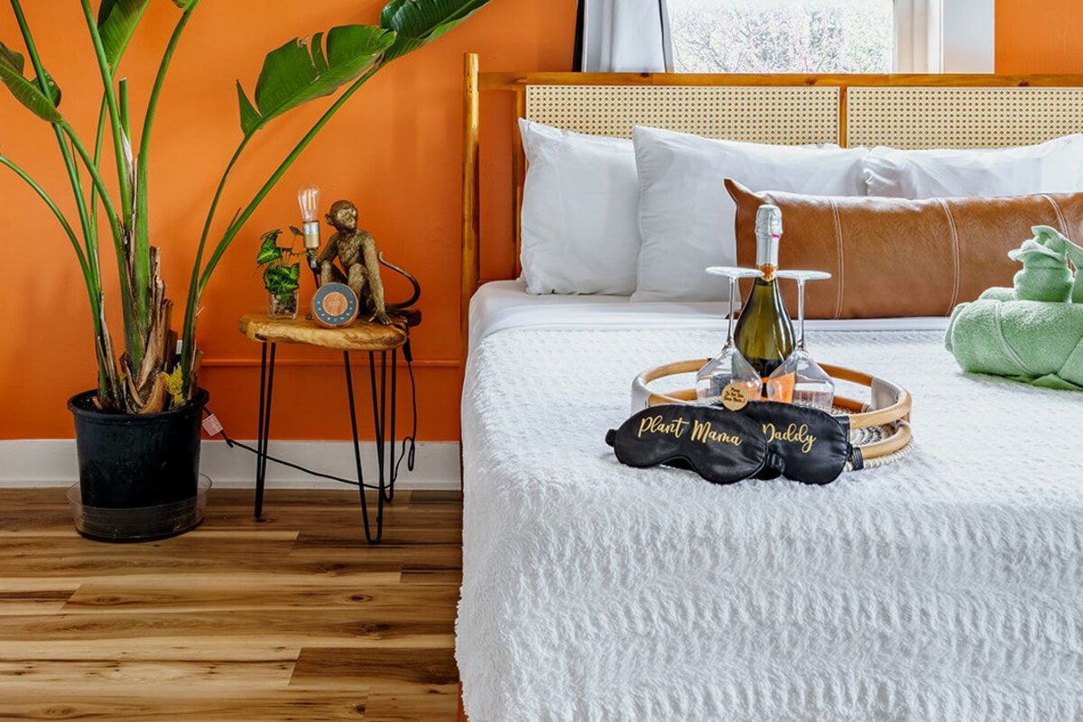 Un dormitorio con una cama de madera, una ventana con cortinas blancas, una planta en maceta y una mesita con una estatua.