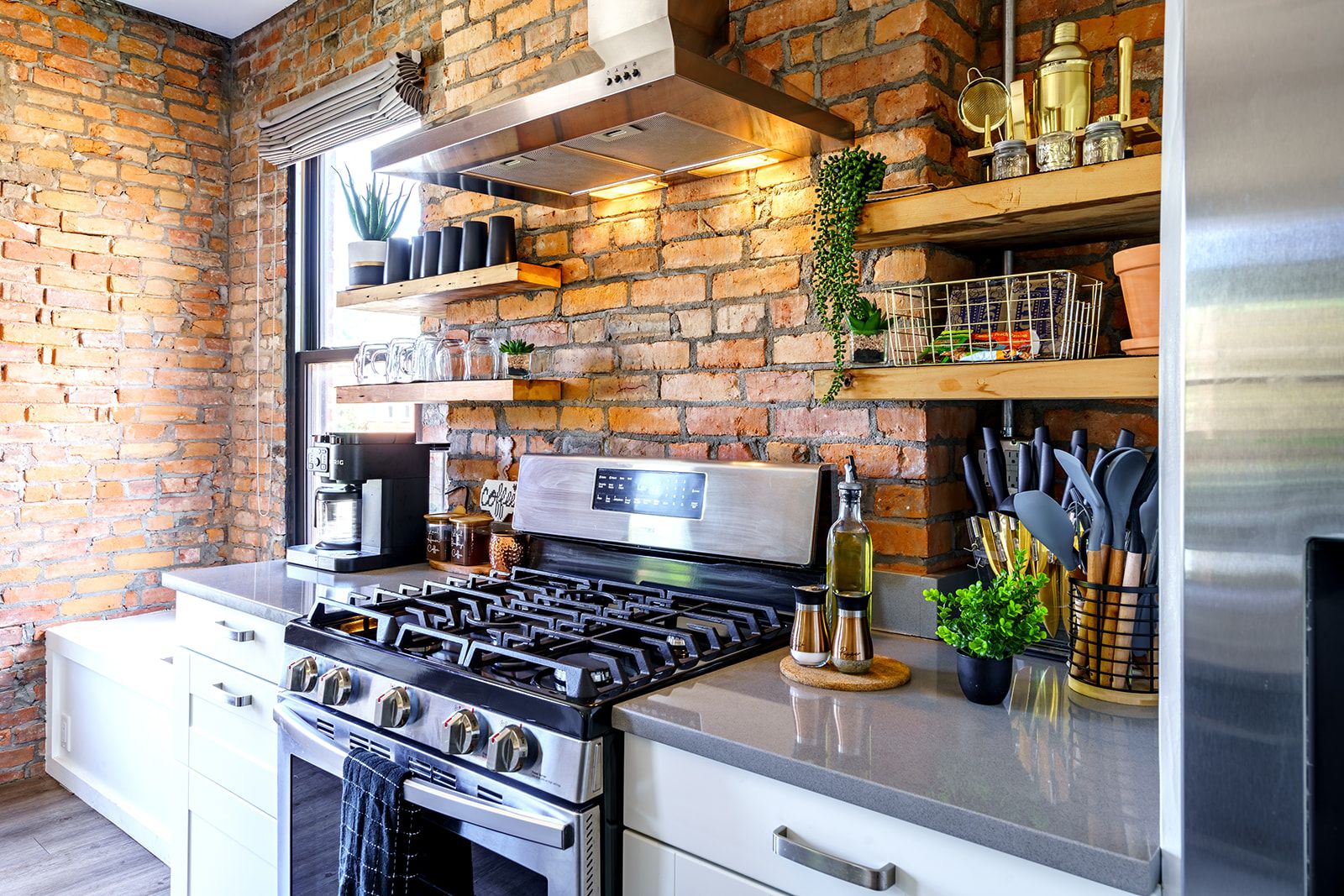 A kitchen with a brick wall, stainless steel appliances, and shelves with various items.
