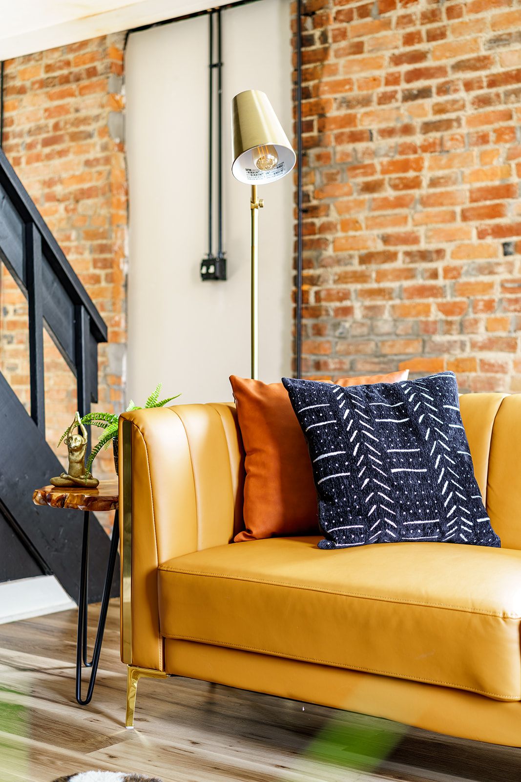 A yellow leather sofa with a patterned pillow, a small plant, and a floor lamp against a brick wall.