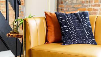 A yellow leather sofa with a patterned pillow, a small plant, and a floor lamp against a brick wall.