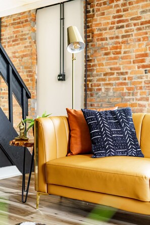 A yellow leather sofa with a patterned pillow, a small plant, and a floor lamp against a brick wall. - Jungle House Downtown Columbus – Modern Boutique Suites (Columbus)