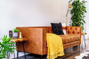 A brown leather sofa with a yellow throw blanket, a small side table with a plant, and a tall floor lamp. - Jungle House Downtown Columbus – Modern Boutique Suites (Columbus)