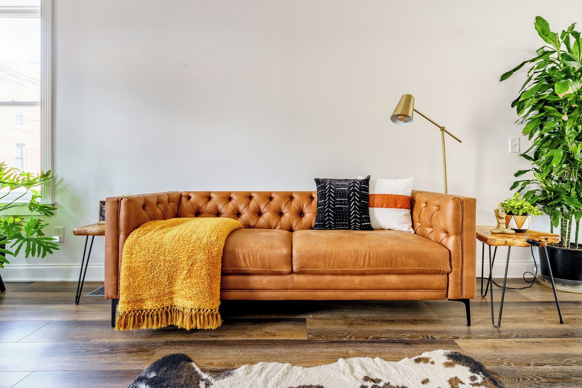 A brown leather sofa with a yellow throw blanket, a black and white patterned pillow, and a gold floor lamp in a room with wooden flooring and green plants.