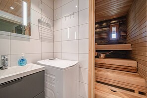 Un baño moderno con baldosas blancas, lavadora empotrada y una sauna de madera visible a través de una puerta abierta.