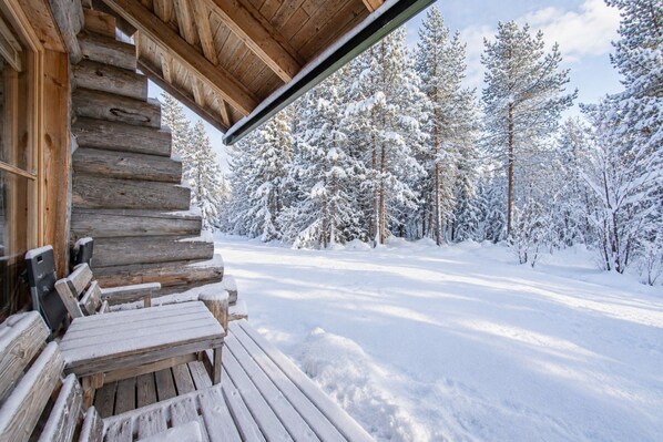Une cabane en rondins avec une terrasse en bois et des bancs, entourée d’une forêt enneigée.