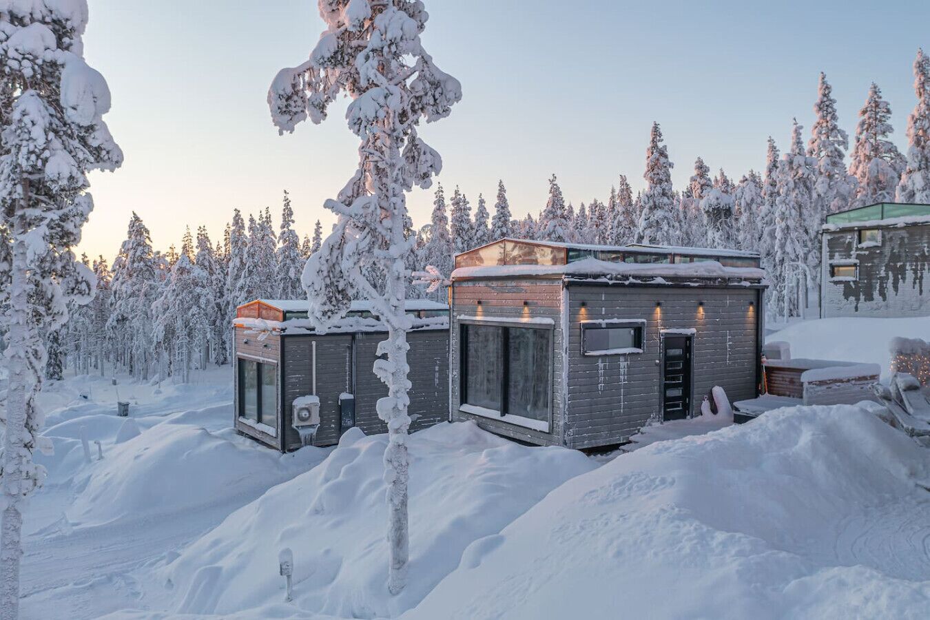 A snow-covered cabin with a small attached structure, surrounded by a forest.