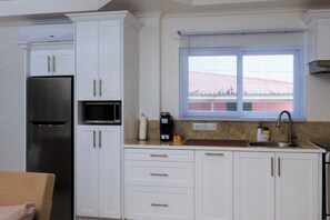 A compact kitchen with white cabinets, a built-in microwave, a stainless steel refrigerator, a window with blinds, and a sink.
