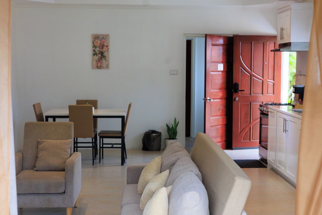 A living room with a sofa, chairs, a dining table, and a kitchen area with cabinets and a red door.