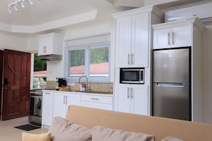 A modern kitchen with white cabinets, a stainless steel refrigerator, a microwave, and a window with blinds. - Agape Suites (Soufrière)